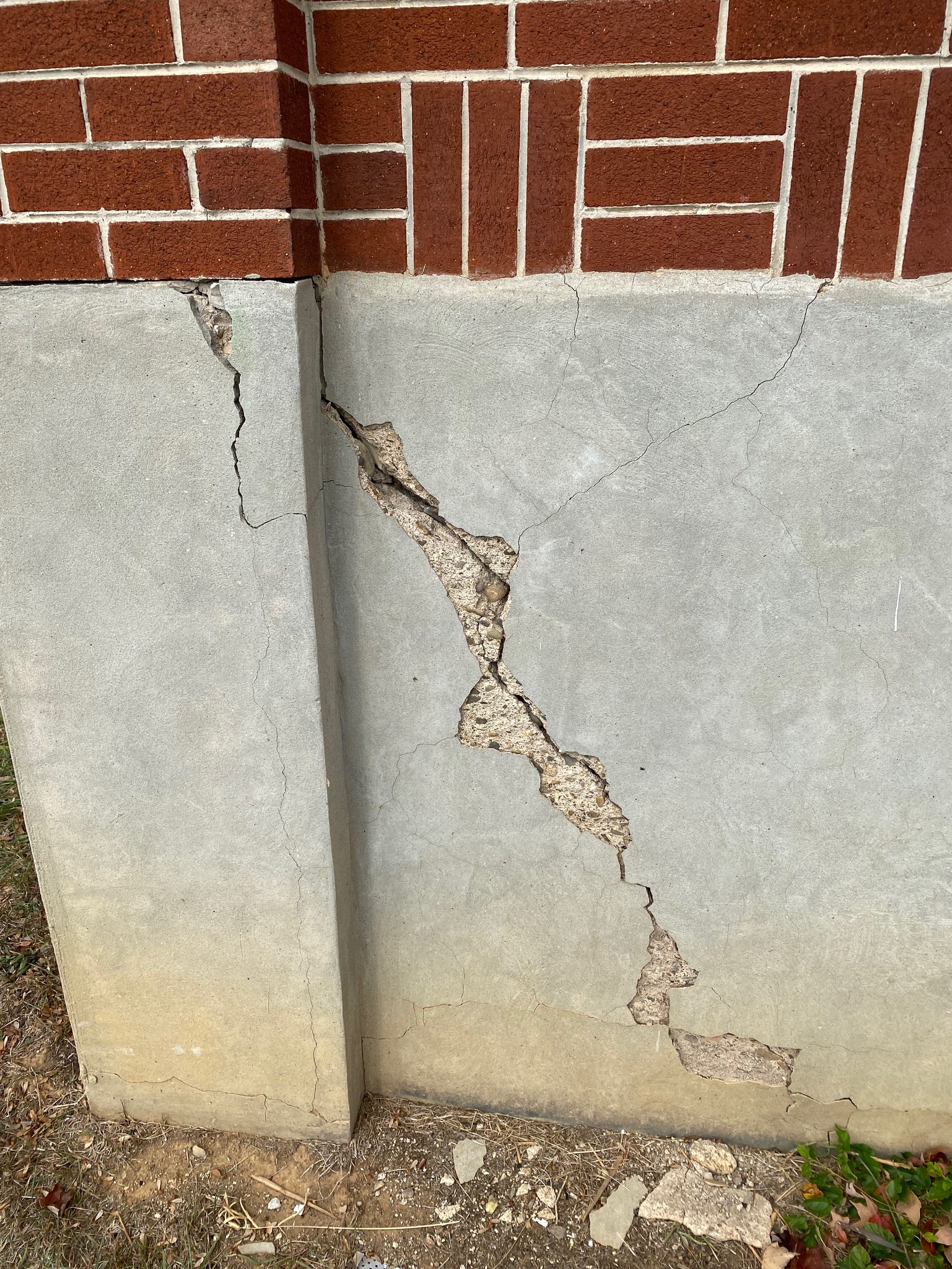 Cracked concrete foundation below red brick wall, showing severe vertical and diagonal fissures. Exposed earth and small debris at the base, suggesting structural issues.