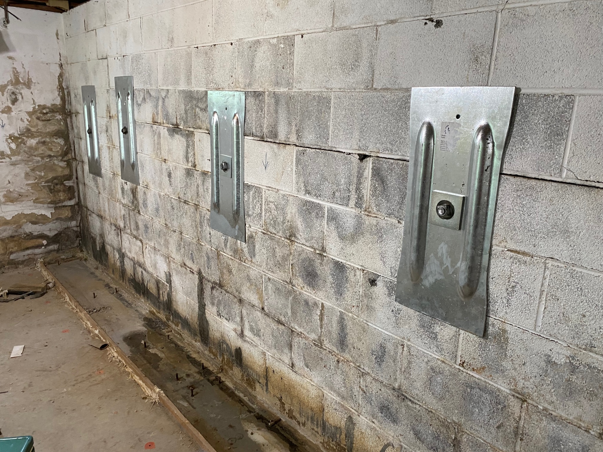 Cinder block basement wall with metal braces installed for reinforcement. The wall shows signs of moisture damage, visible in stained and peeling areas.