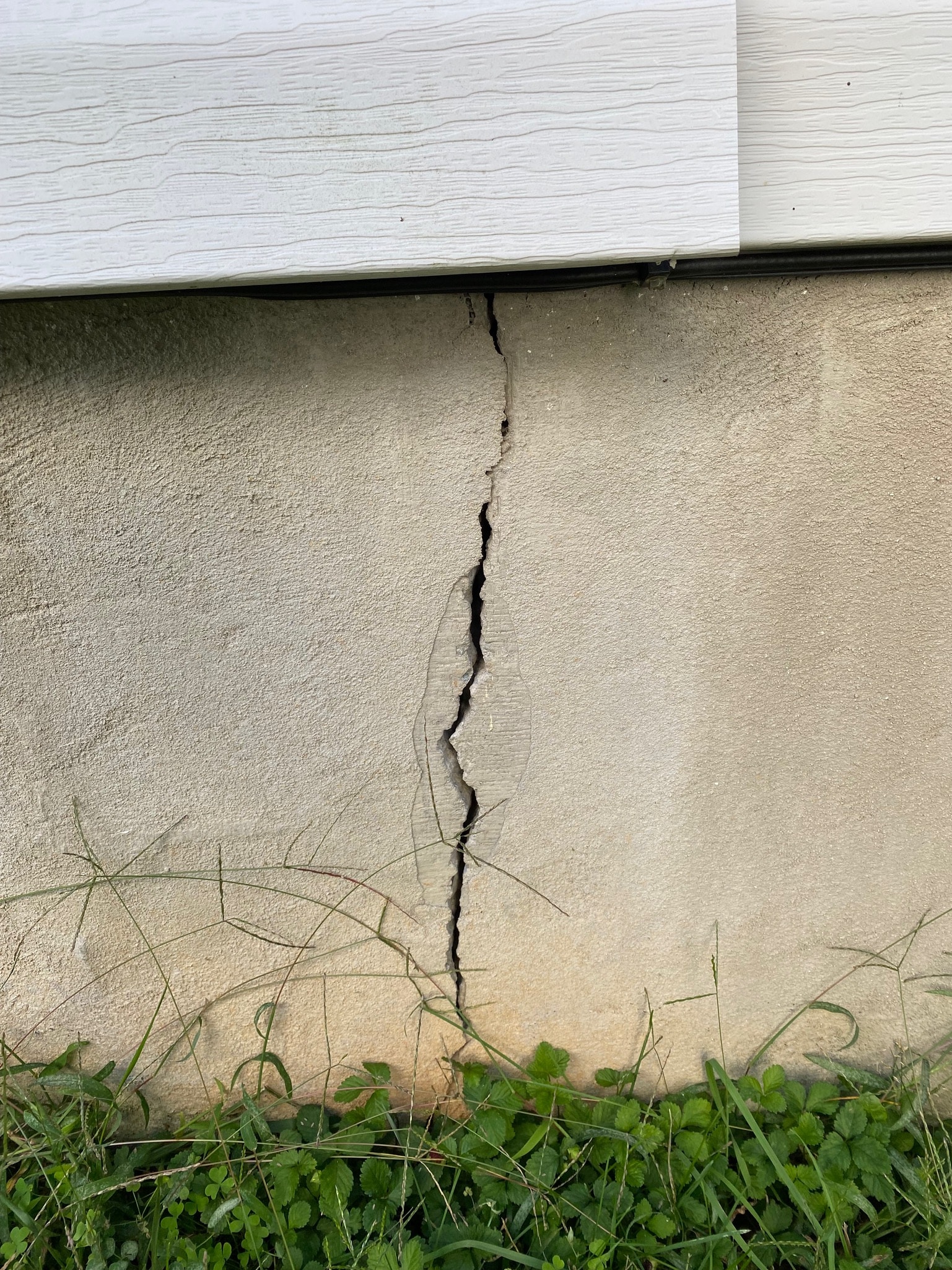 A vertical crack runs through a beige concrete foundation beneath white siding, surrounded by grass. The scene suggests structural damage.