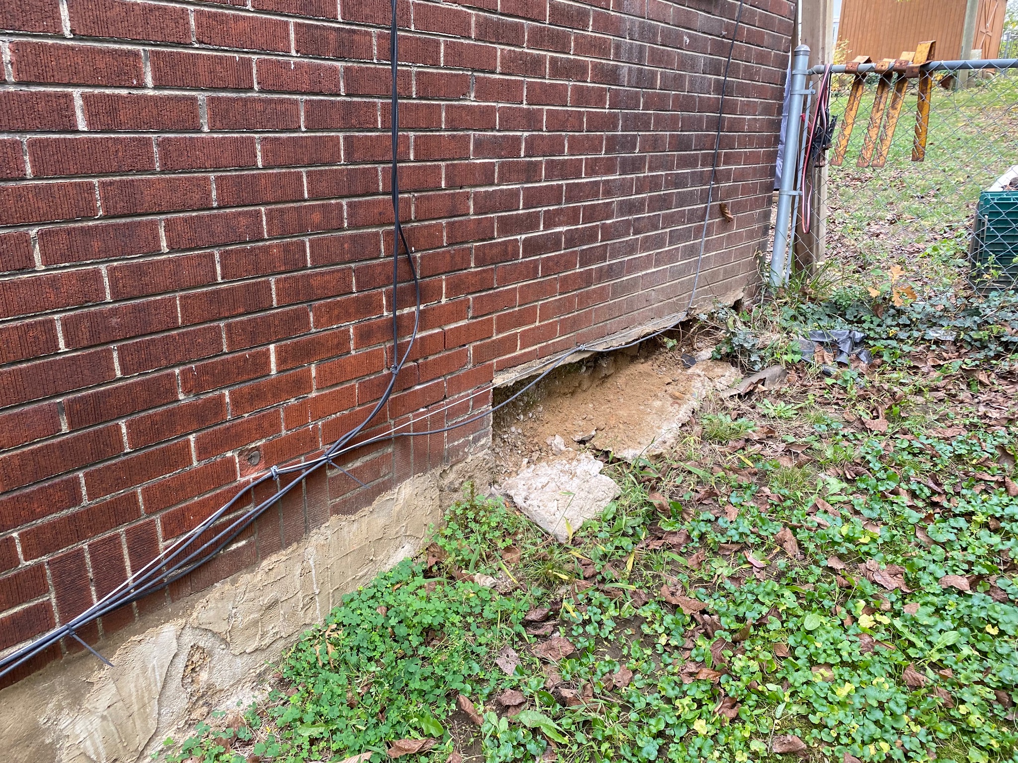 Red brick wall with a large hole near the base, exposing dirt and erosion. Green grass and fallen leaves surround the area, next to a metal fence.