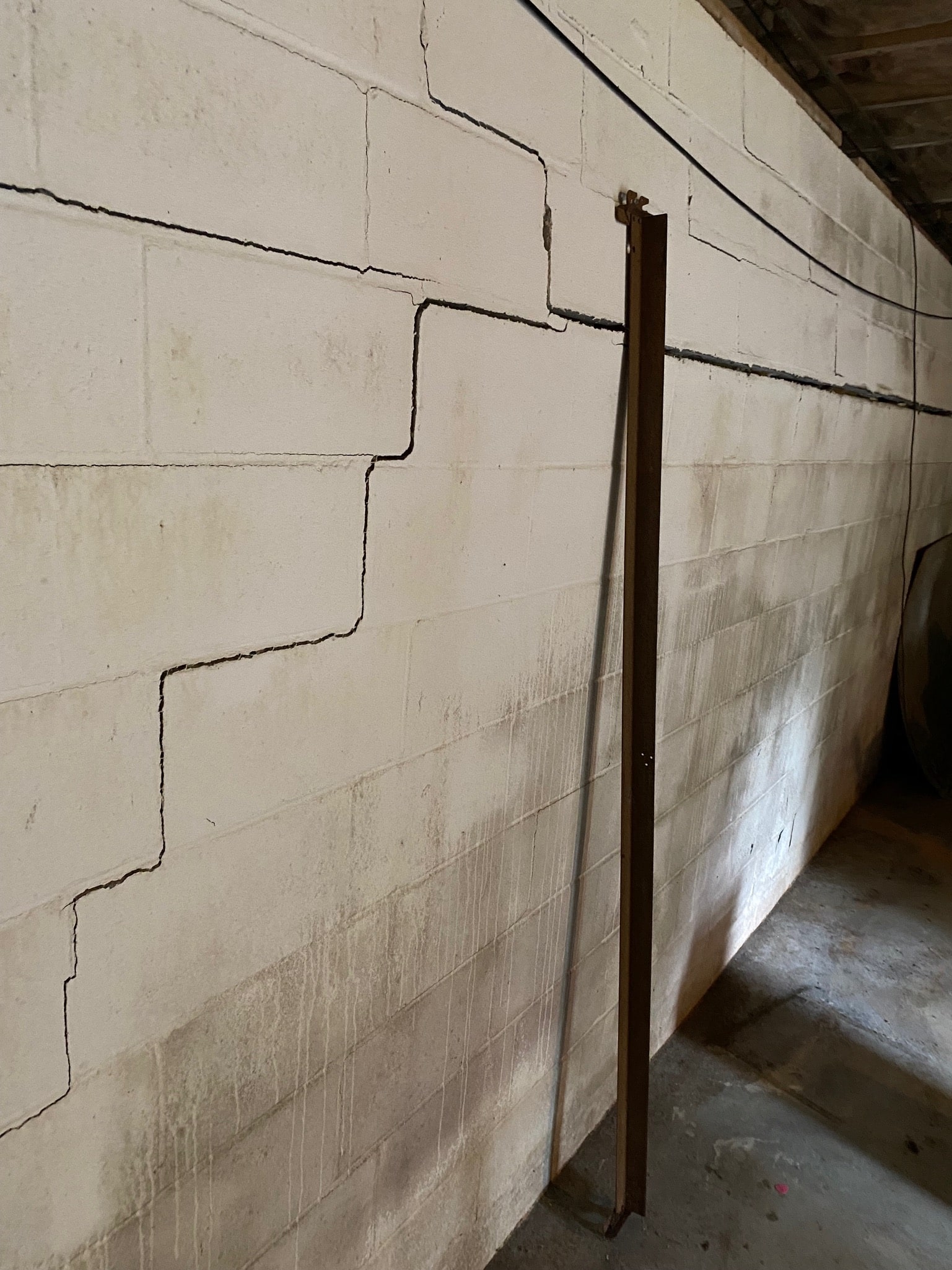Cracked white cinder block wall with visible signs of wear and moisture damage. A metal beam is braced against the wall, indicating structural support.