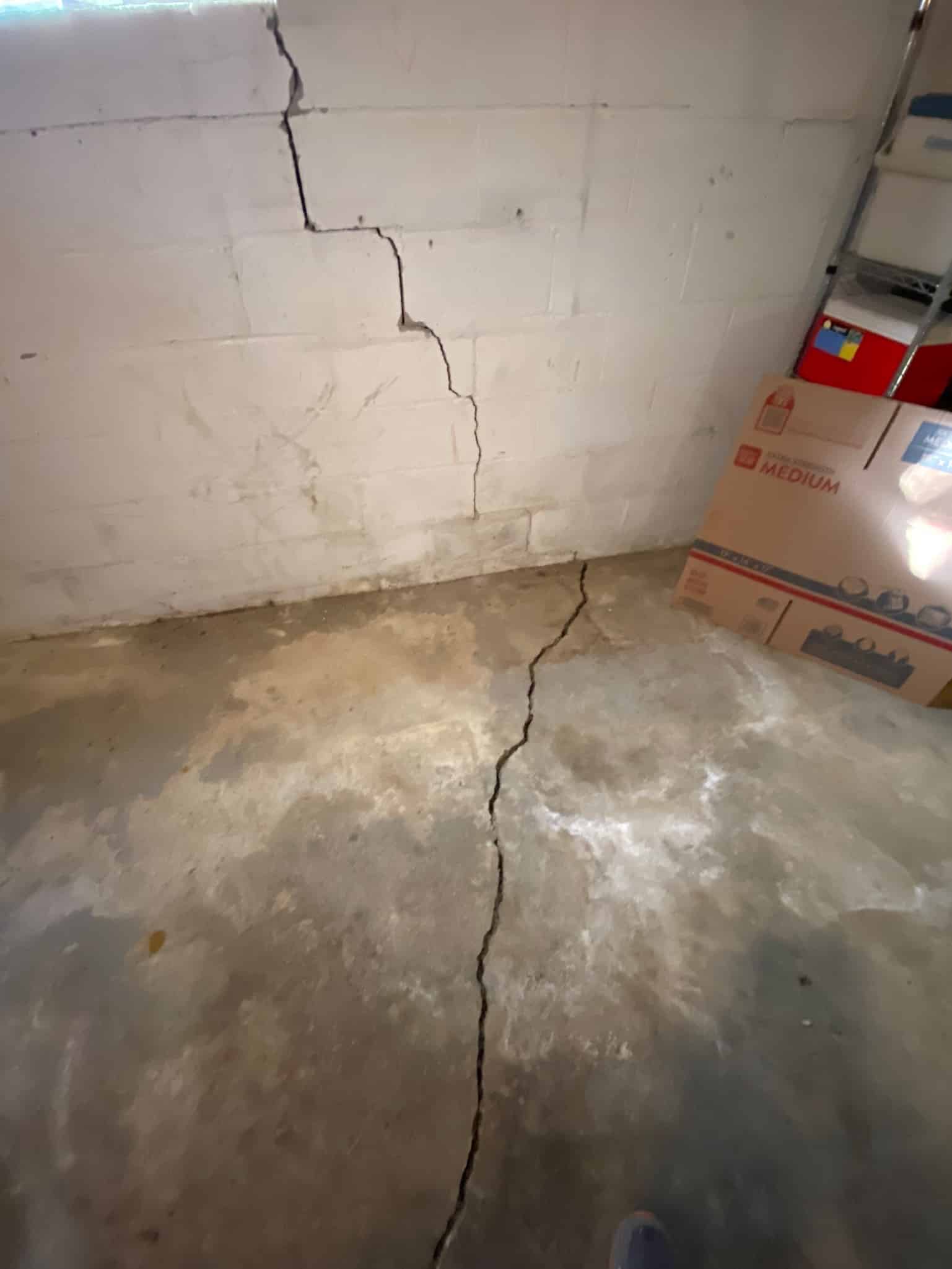 Cracked concrete wall and floor in a basement, with visible horizontal and vertical splits. Cardboard boxes and metal shelving are on the right side.