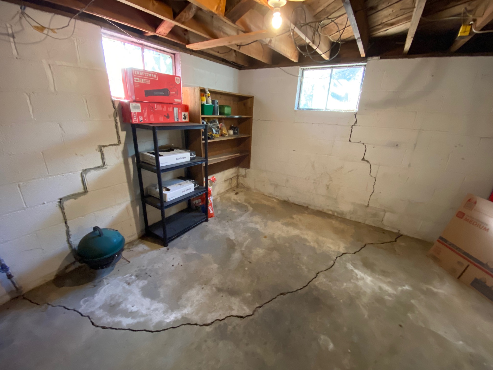A dimly lit basement with cracked cinder block walls and concrete floor. Shelving holds boxes and tools under wooden rafters and small windows.