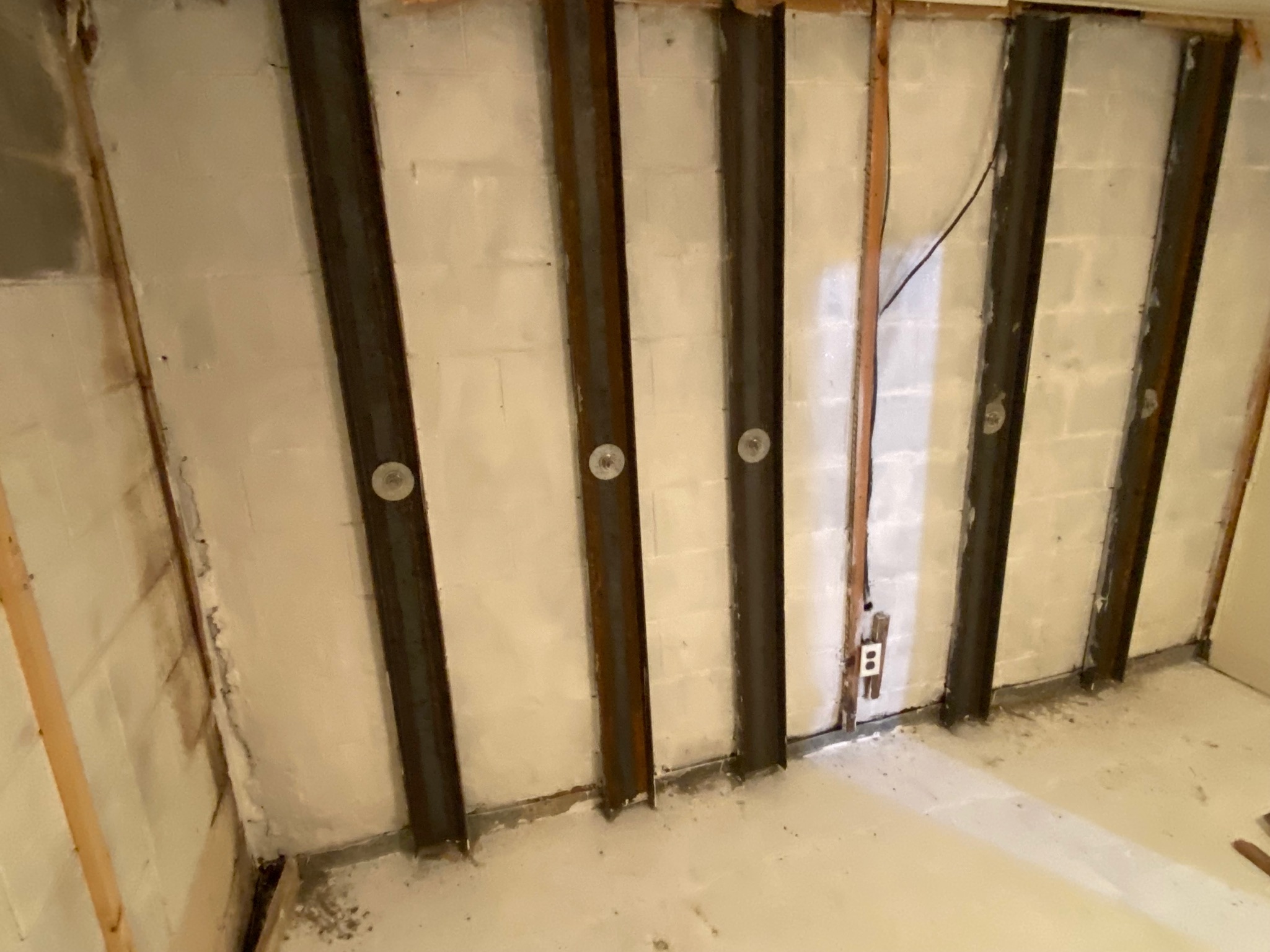 A basement wall with vertical steel beams reinforcing the structure. The concrete blocks are visible and an electrical outlet is mounted on the wall.